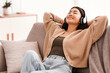 © Prostock-studio - Enjoy The Moment. Smiling asian woman lying on the sofa, leaning back, listening to music, audio book or meditation in wireless headphones in living room, resting and relaxing at home