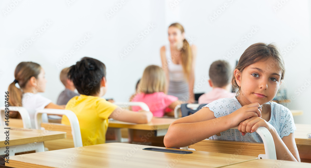 Smiling positive small school girl sitting at school desk in classroom ...