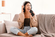 © Prostock-studio - Karaoke App. Portrait of happy asian woman singing her favorite song, using cellphone as mic, wearing wireless headphones. Excited lady sitting on the couch, having fun at home in living room