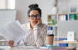 © Prostock-studio - Tired female office worker looking at camera and holding documents, sitting at desk in office. Unhappy woman feeling stressed because of lot of paperwork