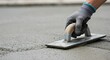 © SimpleDesignStudio - Close-up of a construction worker's gloved hand expertly using a metal trowel to smooth and level freshly poured wet grey concrete on a construction site.