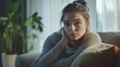 © Letitia - Thoughtful young woman sitting on a couch in a cozy, sunlit room with plants in the background.