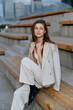 © SHOTPRIME STUDIO - Elegant woman in light blazer and pants sitting on wooden steps outdoors in urban setting with thoughtful expression, fashion style, autumn season