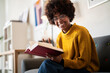 © Studio Marmellata - A young woman with glasses and an afro is sitting on a couch, reading a book. She is wearing a yellow sweater and smiling.