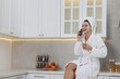© New Africa - Spa day. Beautiful woman with face mask and glass of juice talking on smartphone in kitchen