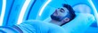 © Tida - Man Relaxing in Blue Light Therapy Chamber - A young man lies peacefully in a blue-lit therapy chamber, eyes closed, enjoying a relaxing treatment