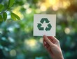 © keke - Hand holding a card with a green recycling symbol against a bright natural background with leaves and sunlight