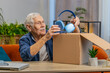 © Andrii Iemelianenko - Happy senior man unpacking delivery parcel with headphones at home. Smiling satisfied grandfather shopper, online shop customer opening cardboard box receiving purchase gift by fast postal shipping