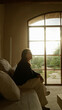 © Krakenimages.com - Young woman relaxing indoors in a mediterranean house with large windows overlooking a garden, seated on a comfortable sofa, enjoying the tranquil and serene ambiance of her home.