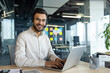 © Liubomir - Portrait of young successful company employee with headset. Online customer support consultant smiling and looking at camera, using laptop for video call, working inside office.
