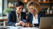 © Nathan - Two young professional women sharing a joyful moment while using a smartphone in a modern office setting.