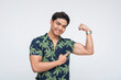© Mdv Edwards - Portrait of a smiling young Southeast Asian man confidently flexing his arm and pointing to his biceps in a tropical shirt. Isolated on a light background.