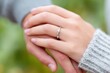 © Natalia - Close-up of two hands intertwined, showcasing a beautiful engagement ring on one hand, with a soft blurred background of greenery, symbolizing love and connection in a romantic setting