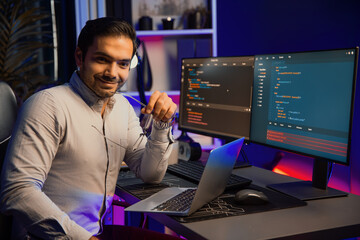 Smiling smart IT developer looking at camera while working on laptop, software coding system at neon light office room, creating application program update latest version online website data. Surmise.