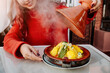 © EdNurg - Tourist enjoying traditional Moroccan food, opening a hot steaming terracotta tajine with vegetables and spices