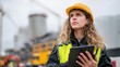 © Attasit - Female engineer wearing a hard hat and high-visibility vest, using a tablet to record data at a construction site, looking directly at the camera.