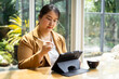 © Leny Studio - Asian Businesswoman A woman is focused on her tablet while seated at a table, surrounded by greenery and natural light, with a cup nearby. Female Digital nomad lifestyle