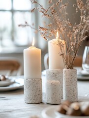 Poster - A tranquil table setting features two lit white candles in textured concrete holders, complemented by a vase of dried beige flowers, creating a serene and minimalist ambiance. : Generative AI
