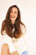 © zvkate - Portrait of a cheerful young woman with long brown hair, wearing a loose white shirt, sitting relaxed and smiling warmly. Natural light studio shot. Happiness, beauty, wellness, lifestyle.