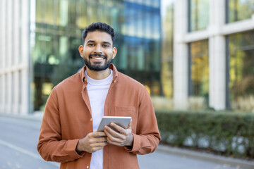 a smiling man stands outdoors, holding a tablet. he wears a casual orange shirt and a white t-shirt,