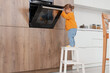 © Pixel-Shot - Cute little girl opening oven on stool in kitchen. Child at risk