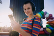 © BullRun - Youthful woman with buzzcut and headphones smiling into camera while holding smartphone, city architecture and colorful sculpture behind, vibrant evening light creates warm modern atmosphere
