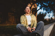 © BullRun - young Hispanic woman sits cross-legged on concrete bench, looking away with thoughtful smile. Dressed in green shirt and jeans, female holds her smartphone, embracing peaceful moment in evening light