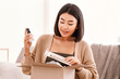 © Prostock-studio - Online Shopping. Smiling asian woman opening parcel box with cosmetics at home, holding makeup products, unpacking gift sitting on the couch at home. Lady satisfied with delivery service