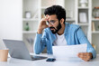 © Prostock-studio - Stressed Young Indian Man Looking At Laptop Screen And Holding Documents, Focused Millennial Eastern Male Cheching Business Reports While Working Remotely At Home Office, Closeup Shot With Free Space