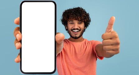 Handsome indian millennial guy showing brand new smartphone with empty white screen and thumb up, smiling at camera over blue studio background, mockup, mobile application concept, panorama