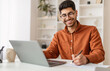© Prostock-studio - Portrait of young smiling Arabic man in glasses using laptop sitting at desk, writing in notebook. Happy guy working remotely, watching webinar training studying online, taking notes signing documents