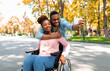 © Prostock-studio - Cheerful black guy hugging his disabled girlfriend in wheelchair, making selfie on smartphone at autumn park. Handicapped couple in love photographing themselves on walk outdoors