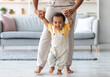 © Prostock-studio - Adorable black infant boy making first steps at home with mother's help. Happy smiling cute african american toddler baby holding mom's hands while walking in living room, closeup with copy space