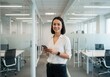 © Over View - Businesswoman using smartphone in modern office hallway, smiling at camera