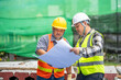 © BESTIMAGE - Two Construction Workers Discussing Blueprints on a Construction Site with Green Safety Nets and Hard Hats in Bright Daylight
