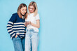 © halayalex - Two young beautiful smiling brunette hipster female in trendy summer clothes. Carefree women posing near blue wall. Positive models having fun. Cheerful and happy. Holding smartphone. Using apps