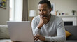 © Vasiliy - African american man in light gray sweater working on laptop with thoughtful expression in cozy living room. Digital technology for remote work, online education and virtual communication platforms