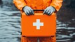 © PStyle - Person in orange protective gear holds a bright orange first aid kit with a white cross near water, suggesting emergency or rescue operations.