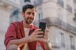 © BullRun - Smiling male tourist checking his phone in a city, possibly using mobile roaming services. warm sunlight and busy streets capture the essence of travel, technology, and digital connectivity on the go