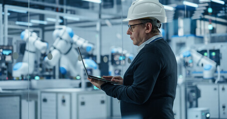 Fototapete - Caucasian Male Engineer in a Hard Hat Using a Laptop Computer to Monitor Robotic Arms in an Advanced Industrial Factory. Specialist Overlooking Modern Automated Electronics Manufacturing