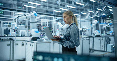 Fototapete - Caucasian Female Engineer Using a Laptop Computer to Monitor Robotic Arms in an Advanced Industrial Factory. Specialist Overlooking Modern Automated Electronics Manufacturing