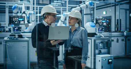 Fototapete - Engineers in a Hard Hats Working in an Industrial Facility, Using Laptop Computer to Monitor Robotic Arms in a Futuristic Factory