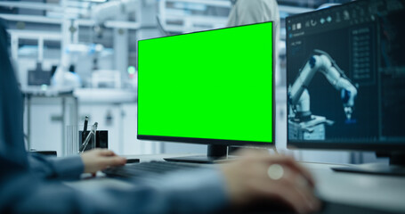 Fototapete - Administrator Working Behind a Desk in a Modern Industrial Facility with Automated Robotic Arms. Anonymous Specialist Working on a Computer with Green Screen Mock Up Template