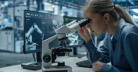 Leinwandbilder - Female Engineer Sitting Behind a Desk in an Electronics Manufacturing Facility, Inspecting High Tech Equipment Under a Microscope and Using Desktop Computer with Dual Screens to Optimize Production