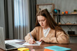 © kinomaster - Young teenage girl studying at home, girl sits at desk with laptop and books, doing homework drawing in notebook with liner and pencil, looking at screen.