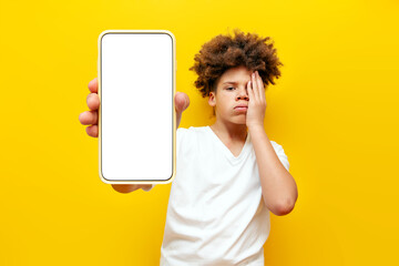 disappointed african american boy showing blank smartphone screen and feeling sad over yellow isolated background, tired curly school boy teenager advertising mobile display and losing and making