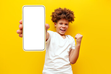 excited african american boy showing blank smartphone screen and celebrating victory on yellow isolated background, curly schoolboy teenager advertising online display and rejoicing in success and