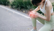 © Jirawatfoto - Asian woman experiencing knee pain during exercise, holding her knee with pained expression, wearing white sports bra and green leggings, on park pathway