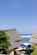 © Natasha Lee - Vertical high angle shot of beach club in Bali with straw roofs next to the ocean with rolling waves on a sunny day with blue skies in Uluwatu, Bali, Indonesia.
