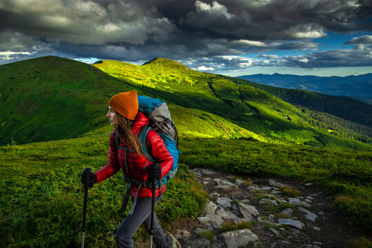 Side view of young woman hiker with trekking poles and backpack climbing stony trail in scenic green mountains under dramatic stormy sky.